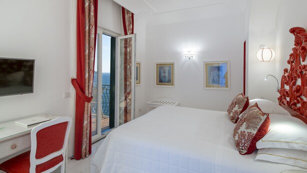 Un dormitorio con cama, escritorio y silla. Hay una ventana con cortinas, un televisor de pared y una puerta que da a un balcón.