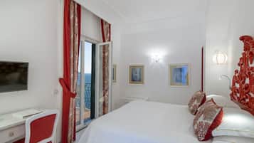 Ein Schlafzimmer mit einem Bett, einem Schreibtisch und einem Stuhl. Es gibt ein Fenster mit Vorhängen, einen an der Wand befestigten Fernseher und eine Tür, die auf einen Balkon führt.