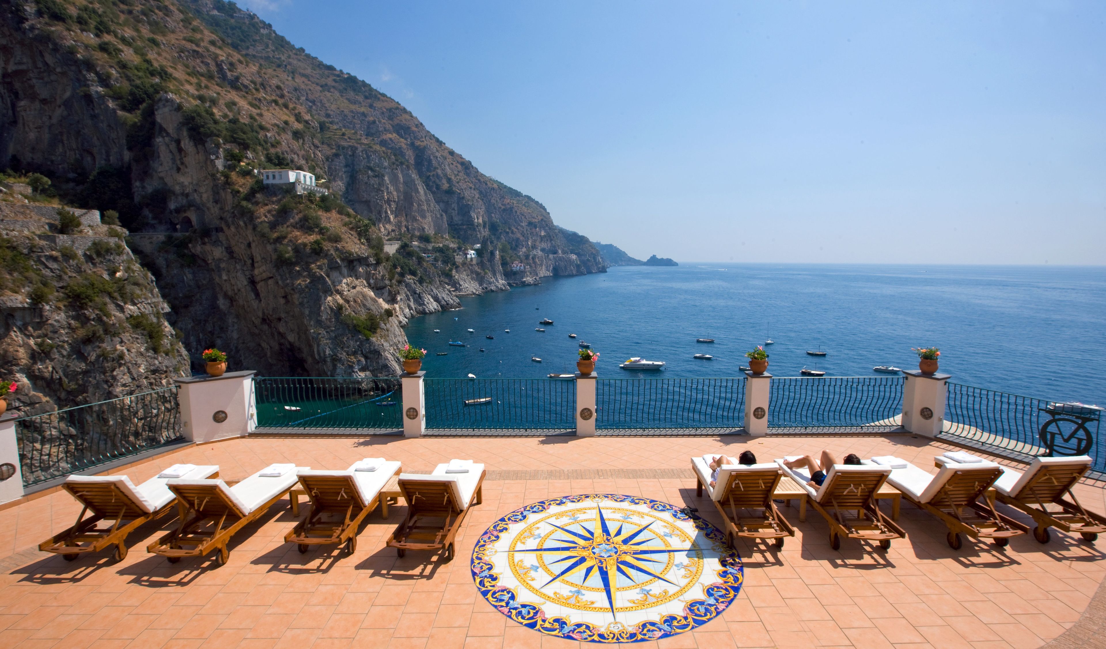 A terrace with sun loungers overlooking a coastal sea with boats, a rocky cliff, and a clear sky.