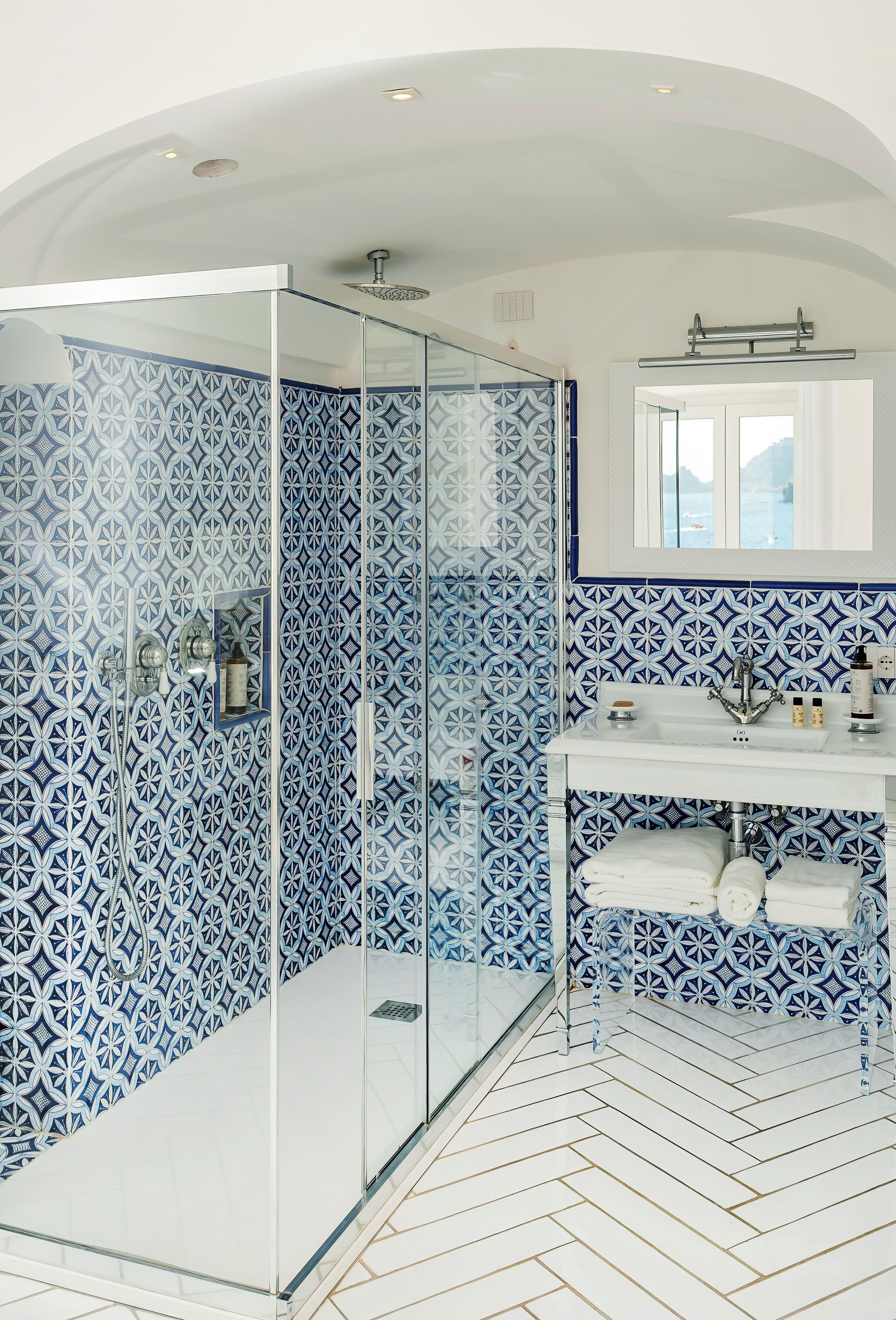 A modern bathroom with a glass-enclosed shower, patterned blue and white tiles, a white sink with a mirror above, and a herringbone floor pattern.