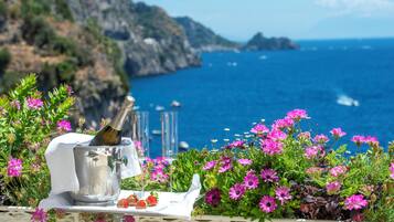 Una mesa con una botella de champán, copas, fresas y flores rosadas, con vista al mar y acantilados.