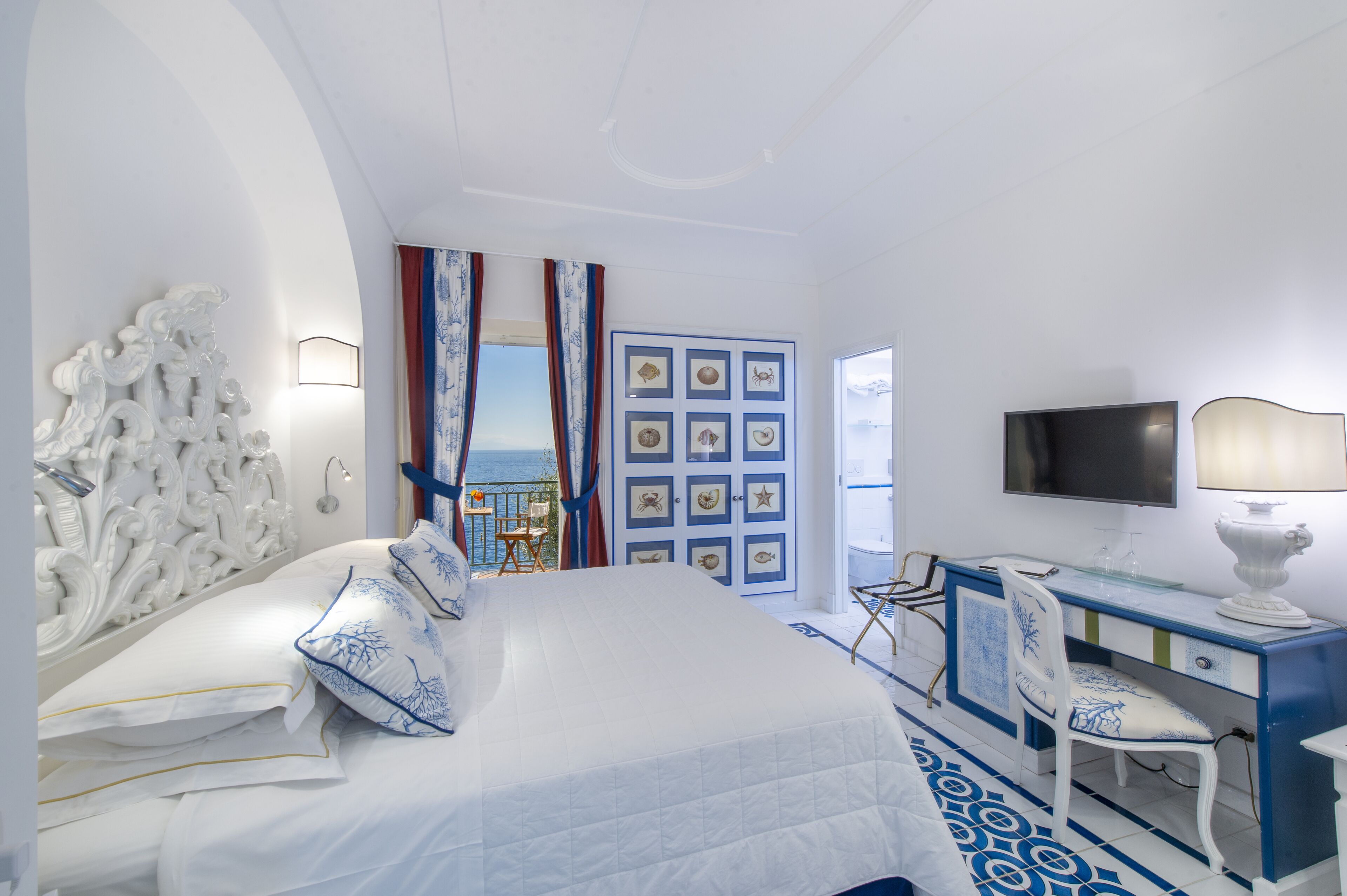 A hotel room with a bed, a desk, a chair, a TV, and a view of the sea.