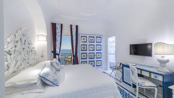 A hotel room with a bed, a desk, a chair, a TV, and a view of the sea.