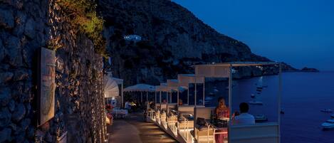 A seaside restaurant with outdoor seating along a rocky cliff, overlooking the sea.