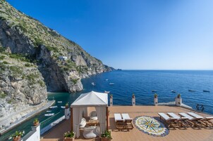 Una terraza con sillones de descanso y un toldo, con vistas a una zona costera con barcos y acantilados.