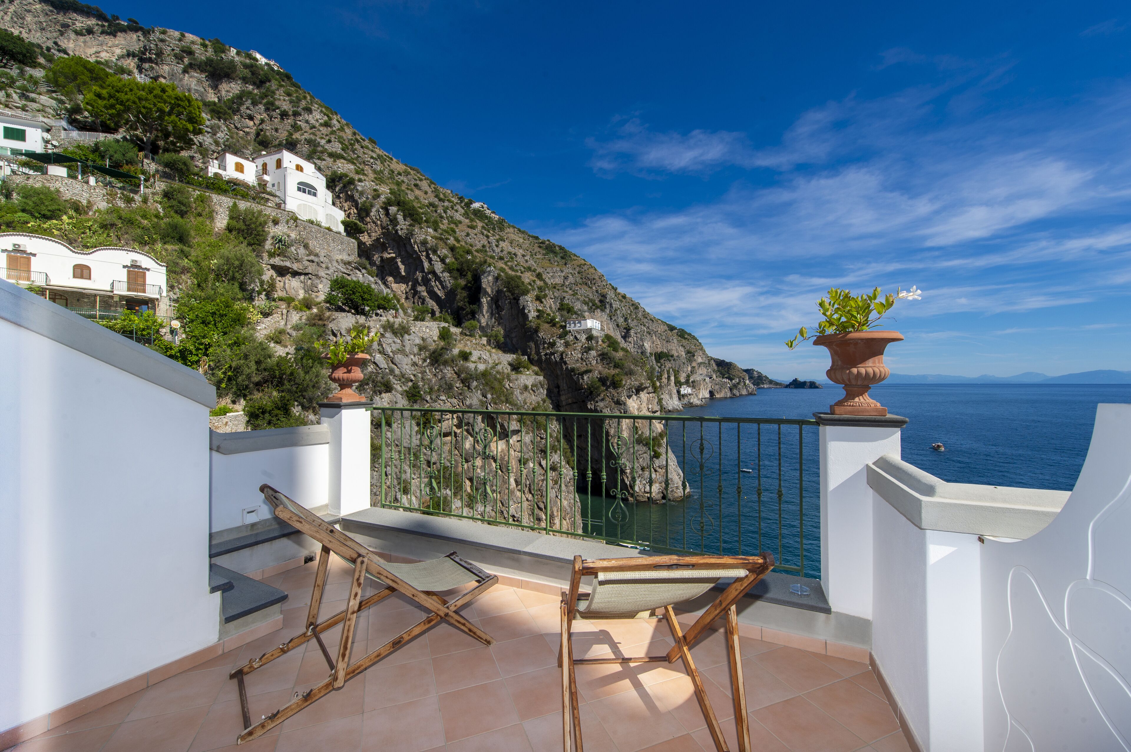 A balcony with two wooden chairs, a potted plant, and a view of the sea and rocky hillside.