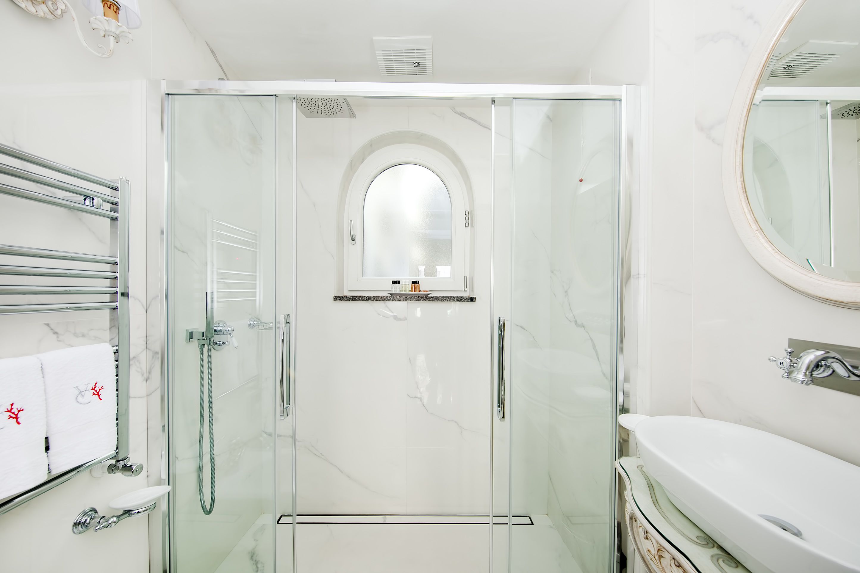 A modern bathroom with a glass-enclosed shower, a marble-tiled wall, a white sink, and a large mirror.