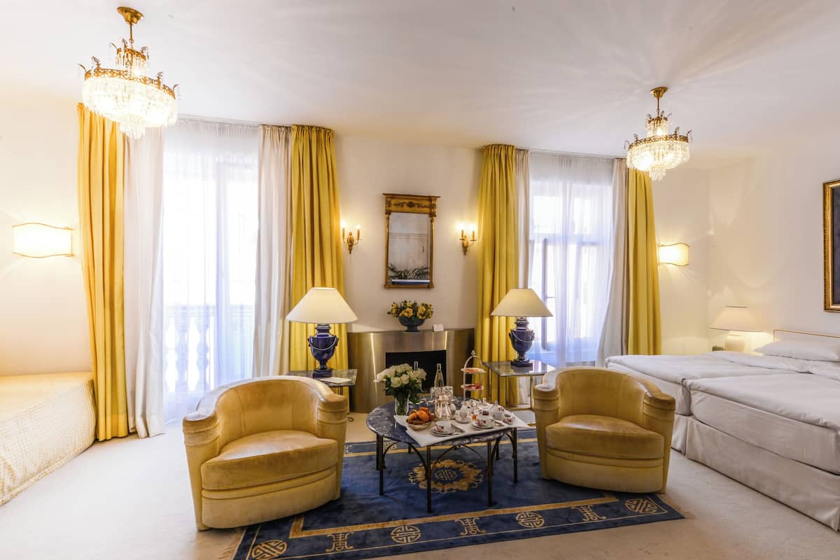 A hotel room with a bed, two armchairs, a small table with a vase of flowers, two lamps, a painting on the wall, and a chandelier.