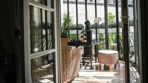 A sunlit balcony with wicker furniture, a small table with a lamp, and a view of greenery outside. - Opera Hotel (Munich)