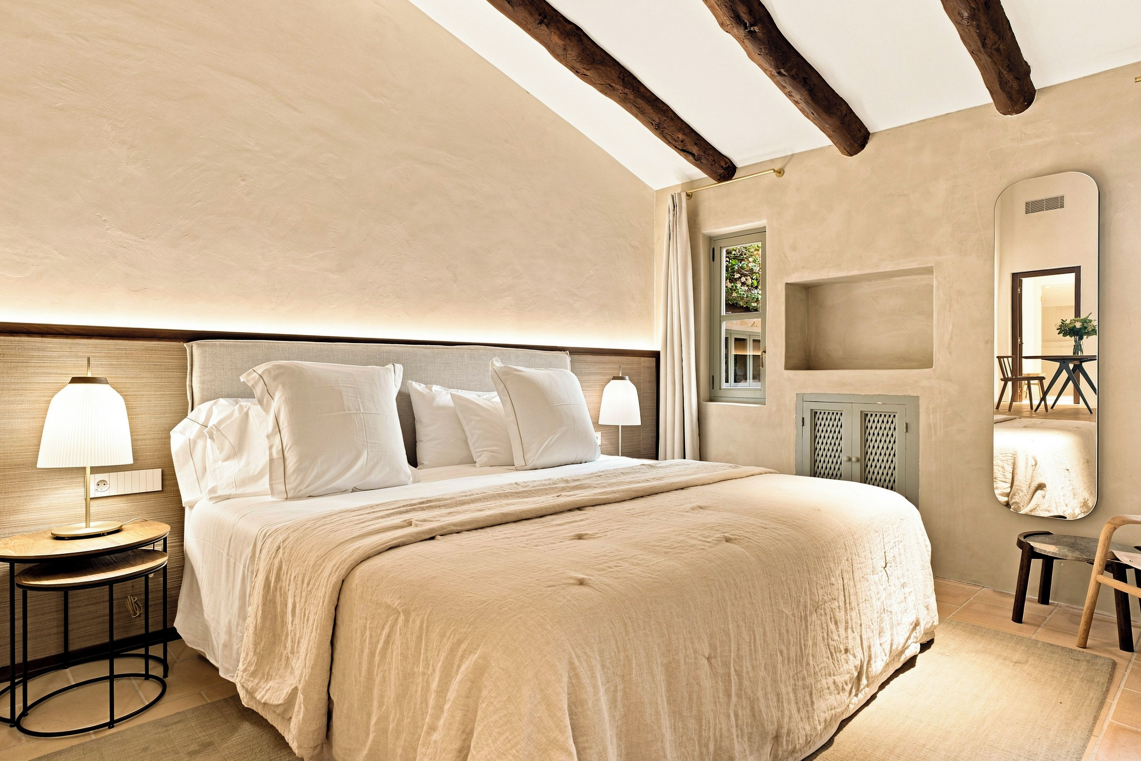 A bedroom with a large bed, two bedside tables with lamps, a wooden headboard, a mirror, and a small table with a chair.