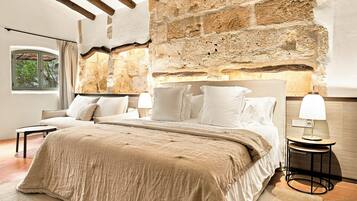 A bedroom with a stone wall, a bed with white bedding, a nightstand with a lamp, and a window with a view of trees.
