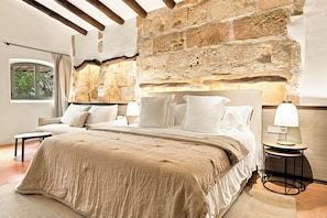 A bedroom with a stone wall, a bed with white bedding, a nightstand with a lamp, and a window with a view of trees. - Rotana la Reserva (Manacor)