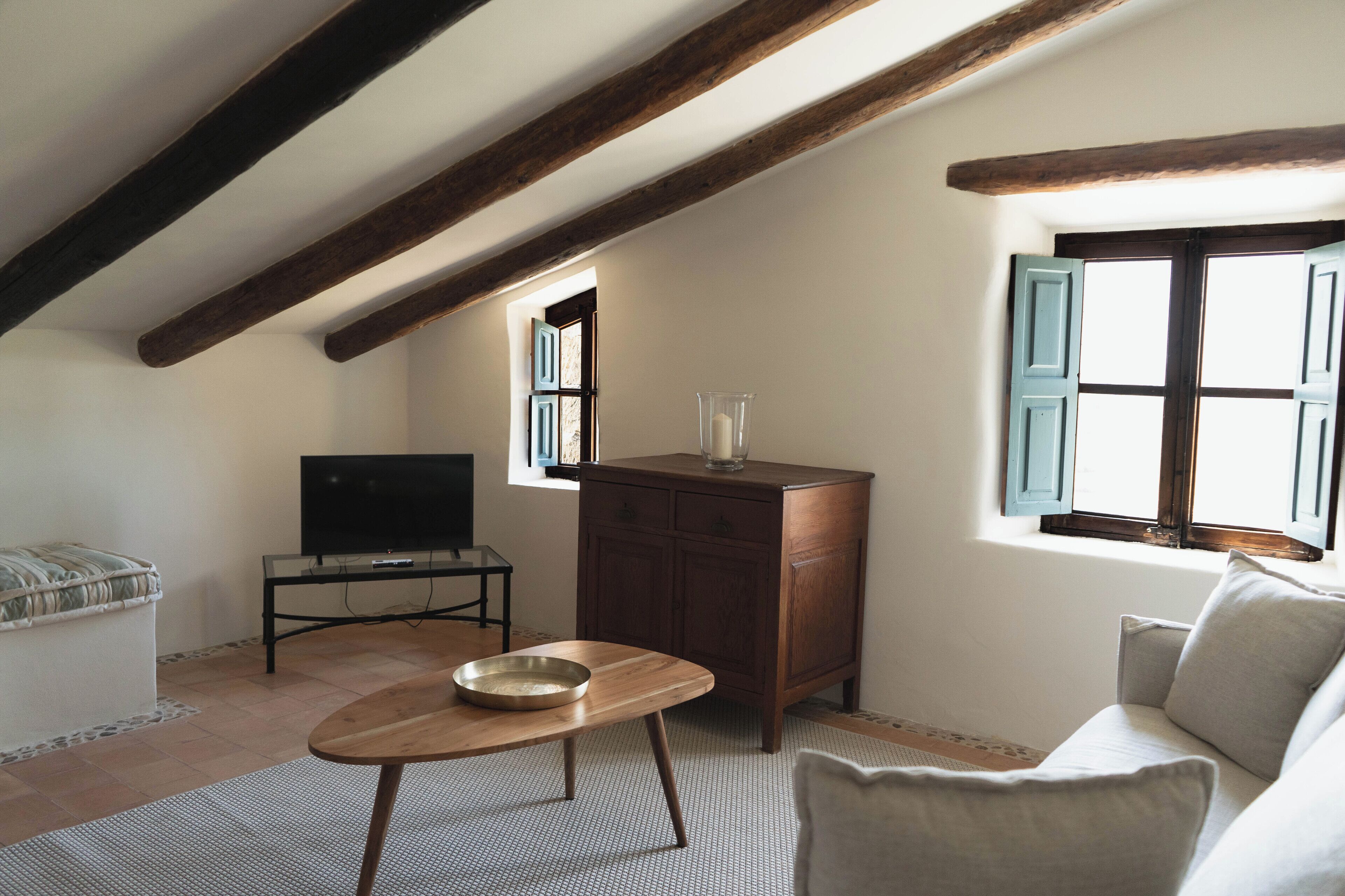 A cozy attic room with exposed wooden beams, a wooden table with a bowl, a sofa, a TV, and a cabinet.