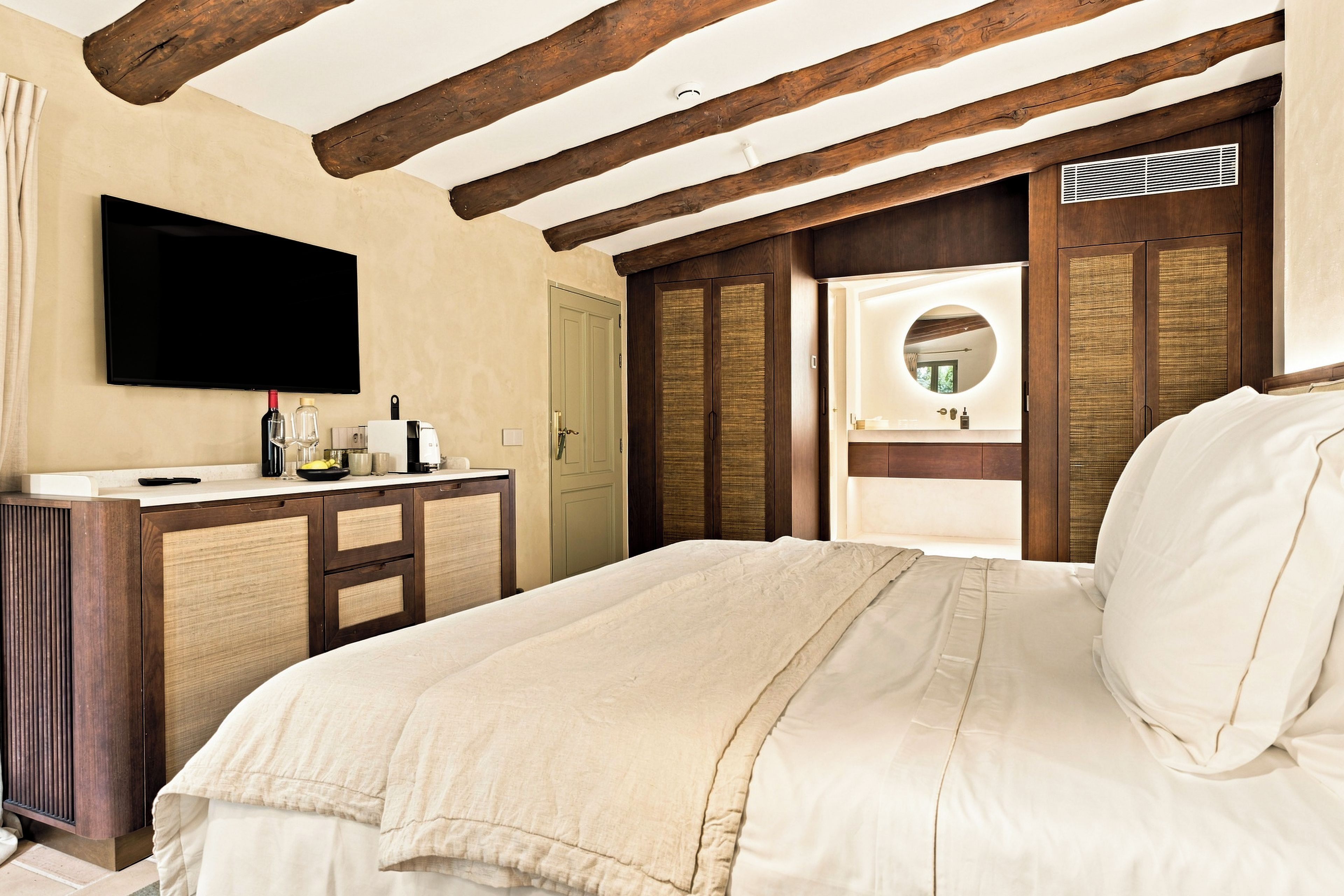 Une chambre d’hôtel avec un grand lit, un téléviseur à écran plat, un bureau avec une bouteille et des verres, et une porte donnant sur une salle de bain équipée d’un miroir.