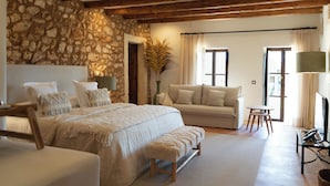 A bedroom with a stone wall, wooden beams, a bed with a bench, a sofa, and a TV. - Rotana la Reserva (Manacor)