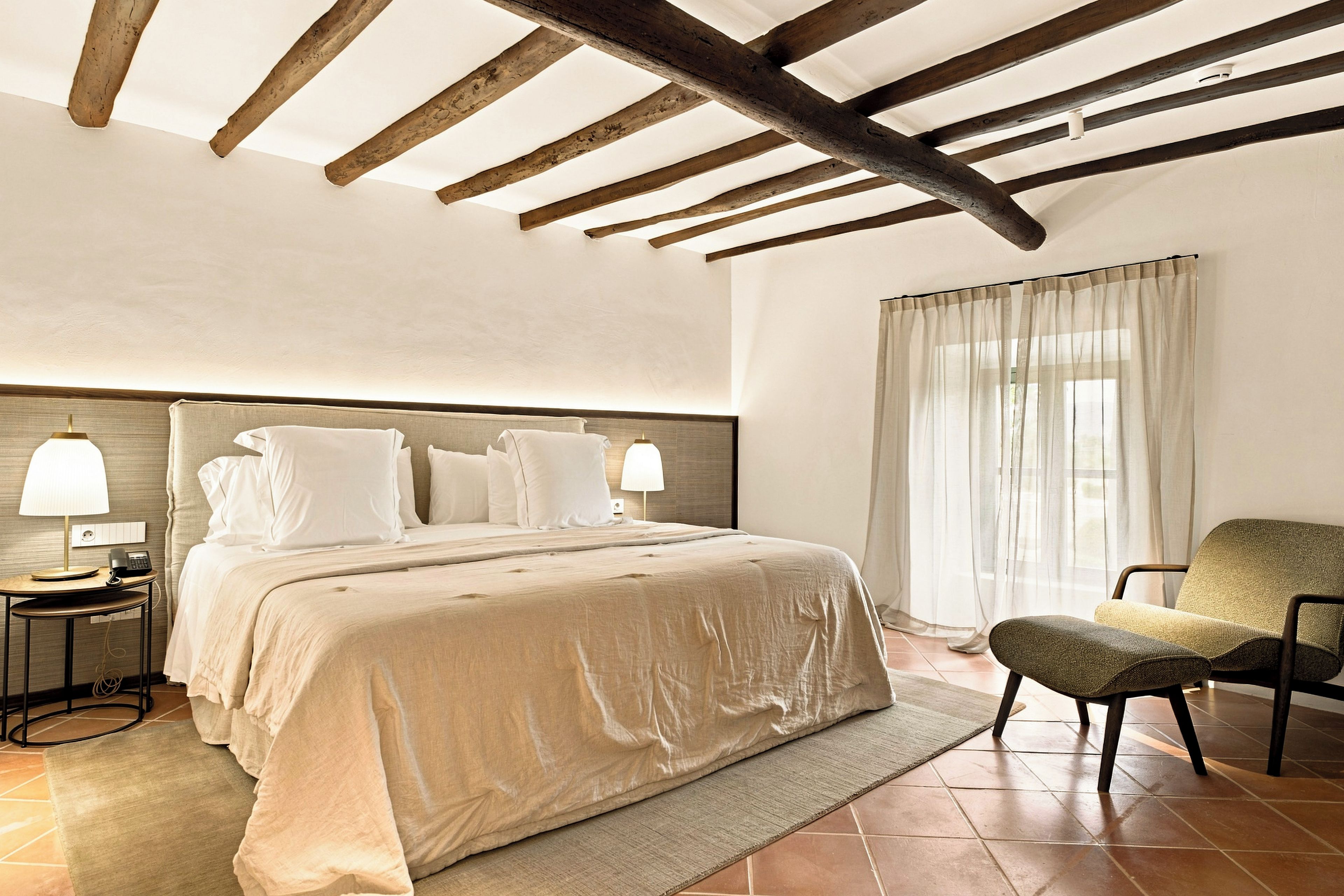 Une chambre à coucher avec un grand lit, deux lampes de chevet, une chaise et une fenêtre avec des rideaux légers.