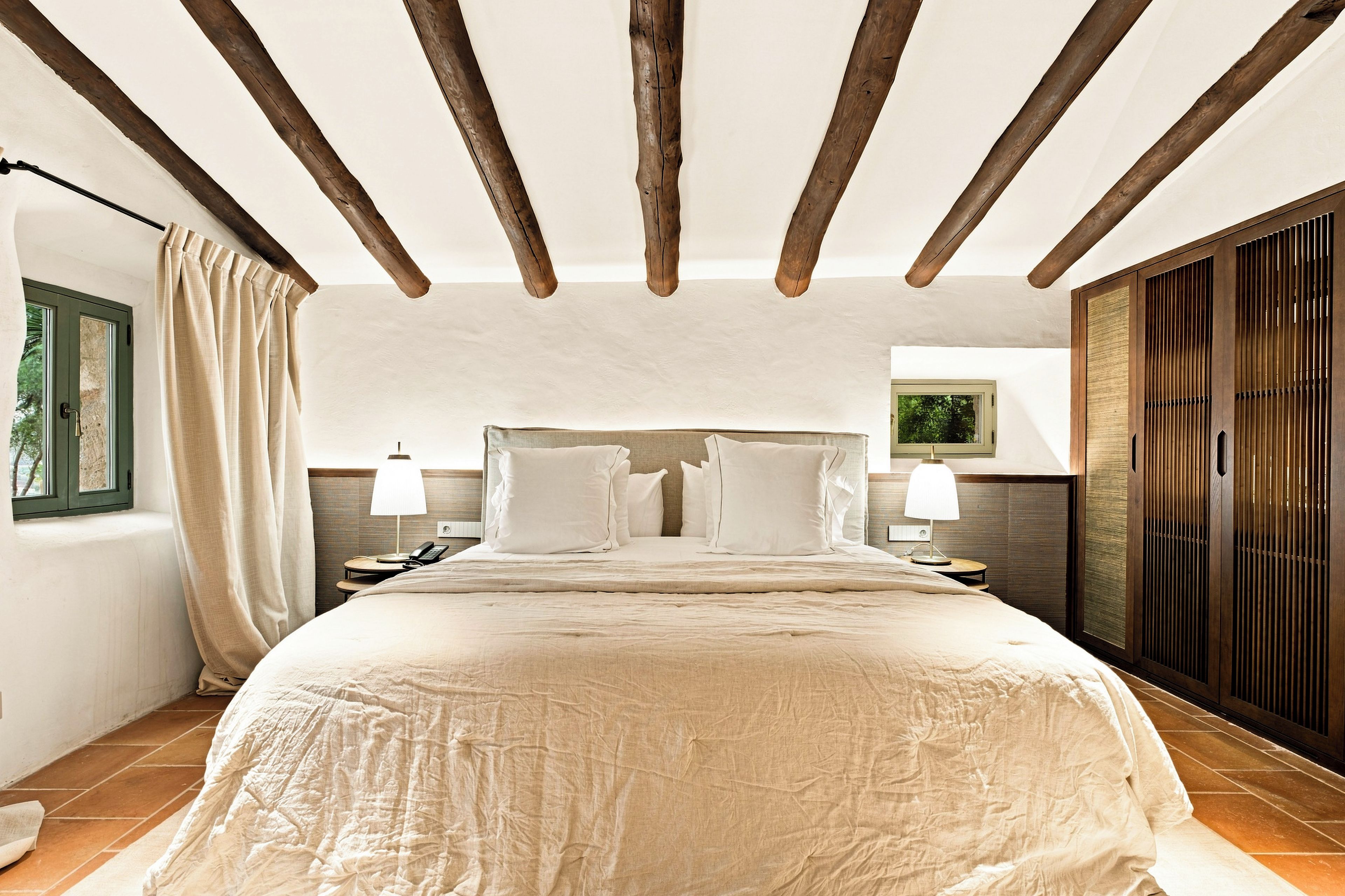 A bedroom with a large bed, wooden beams on the ceiling, a nightstand with a lamp, and a window with curtains.