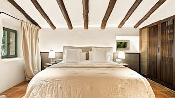 A bedroom with a large bed, wooden beams on the ceiling, a nightstand with a lamp, and a window with curtains.