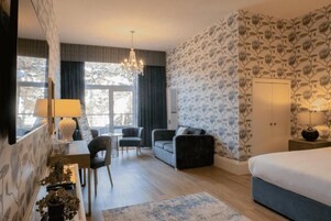 Een hotelkamer met een bed, een bureau met een lamp, een sofa, een kroonluchter en uitzicht op bomen.