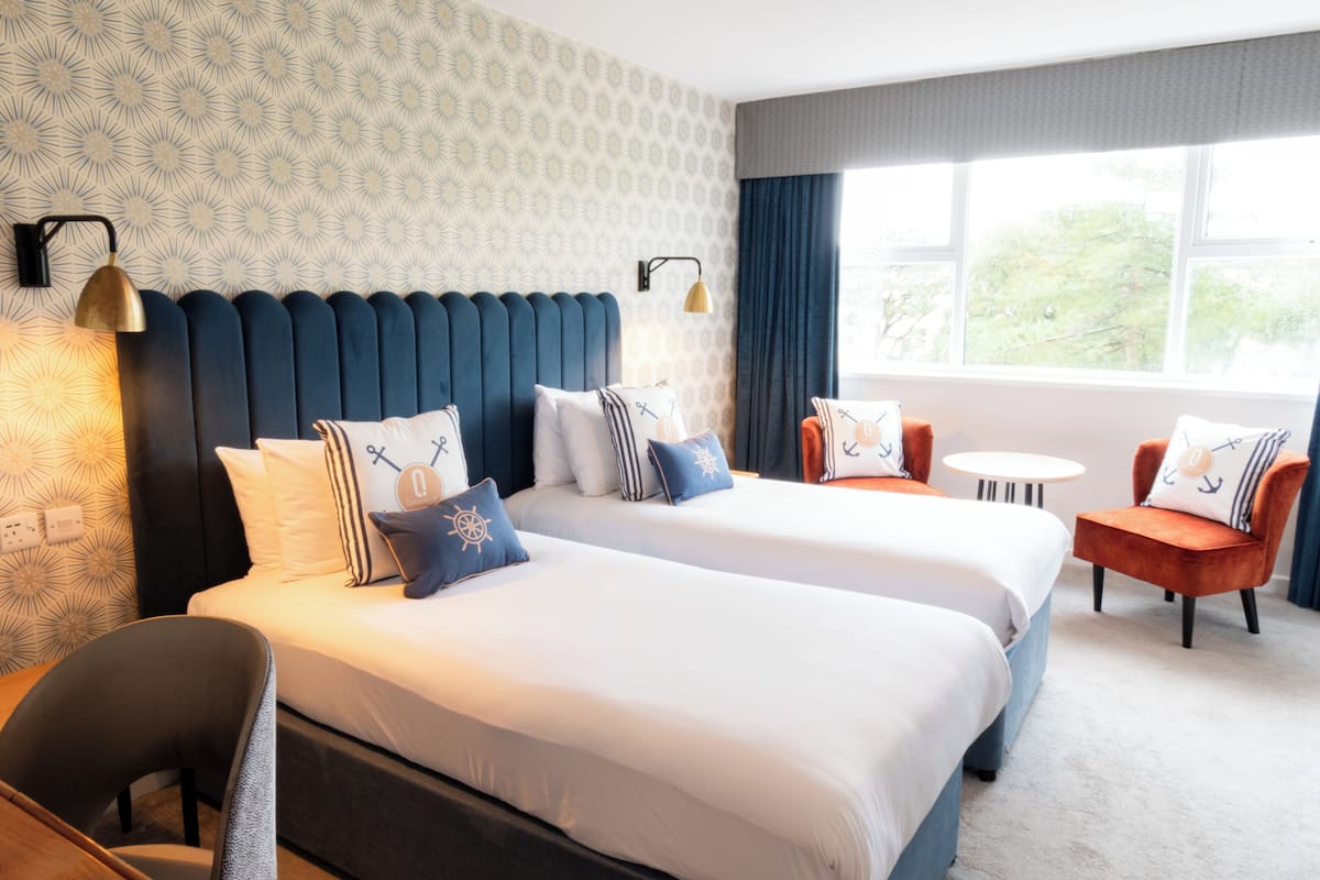 Hotellihuone, jossa on kaksi sänkyä, pieni pöytä, kaksi tuolia ja ikkuna, josta on näkymä.