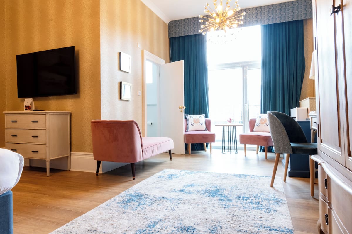 Moderni hotellihuone, jossa on taulutelevisio, vaaleanpunainen sohva, harmaa nojatuoli, puinen lipasto ja suuri ikkuna, jossa on verhot.