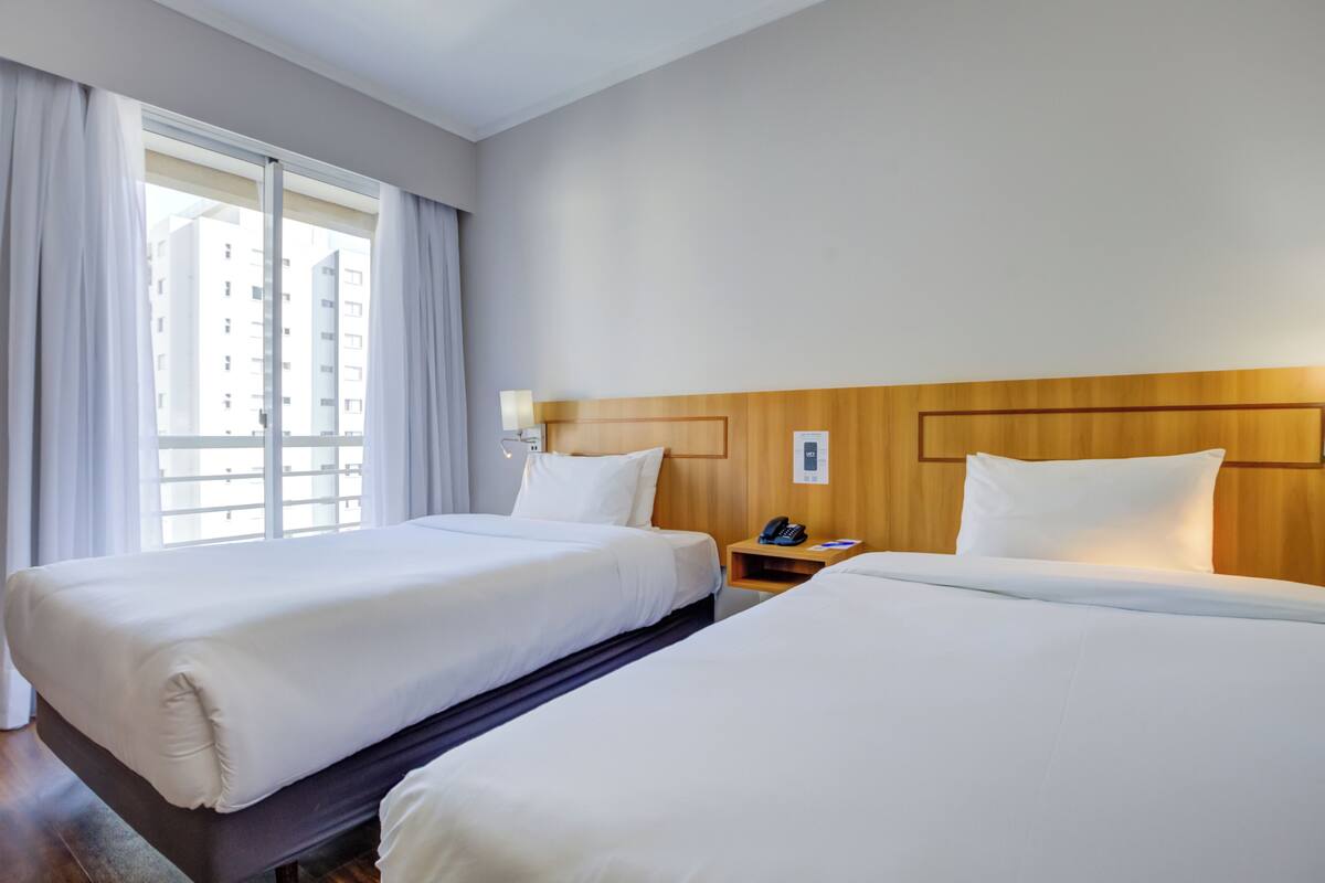 Ein Hotelzimmer mit zwei Betten, einem Holzkopfende, einem Nachttisch mit Telefon und einem Fenster mit Vorhängen.