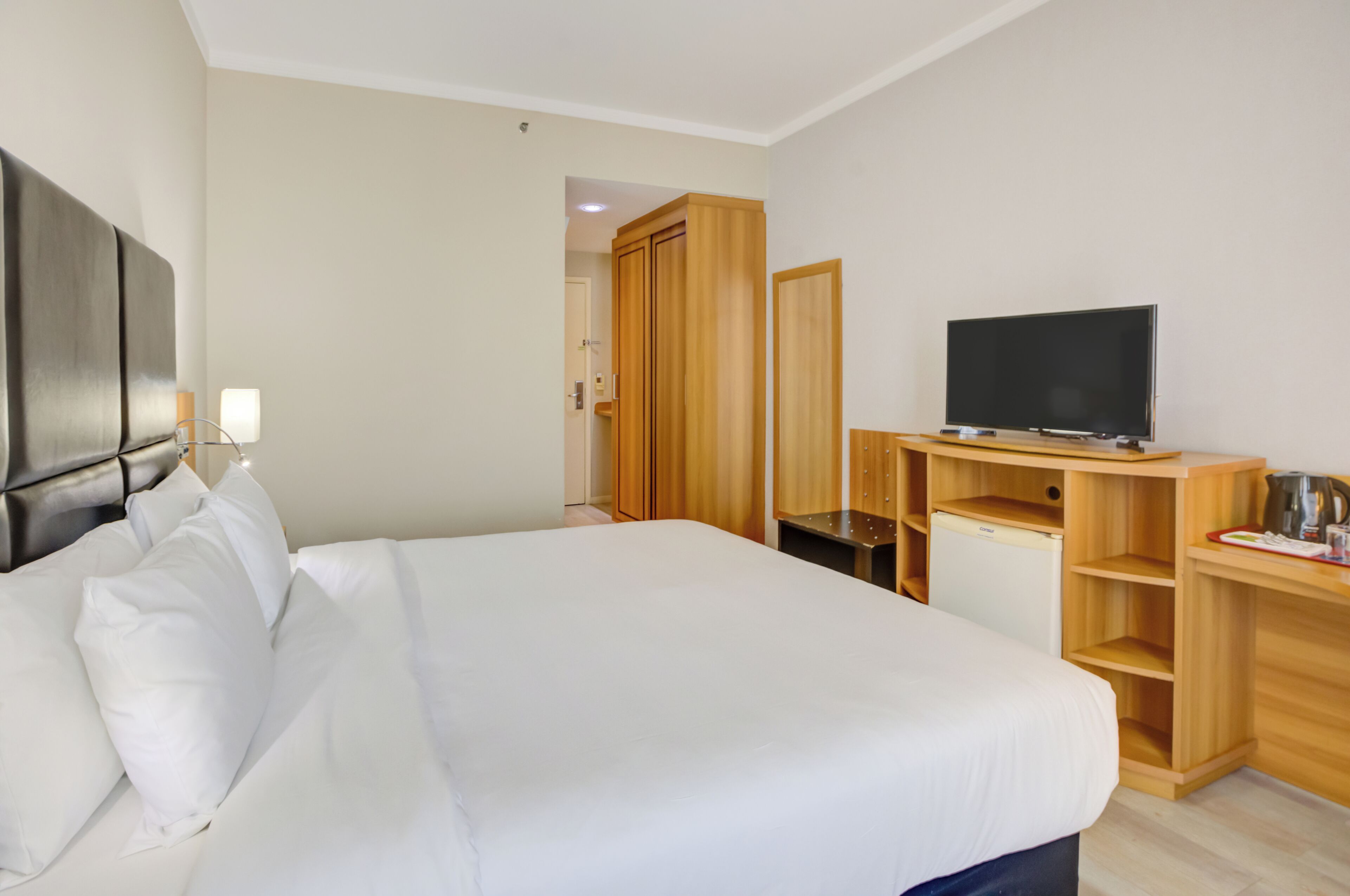 A hotel room with a bed, a television, a wardrobe, and a small desk with a kettle.