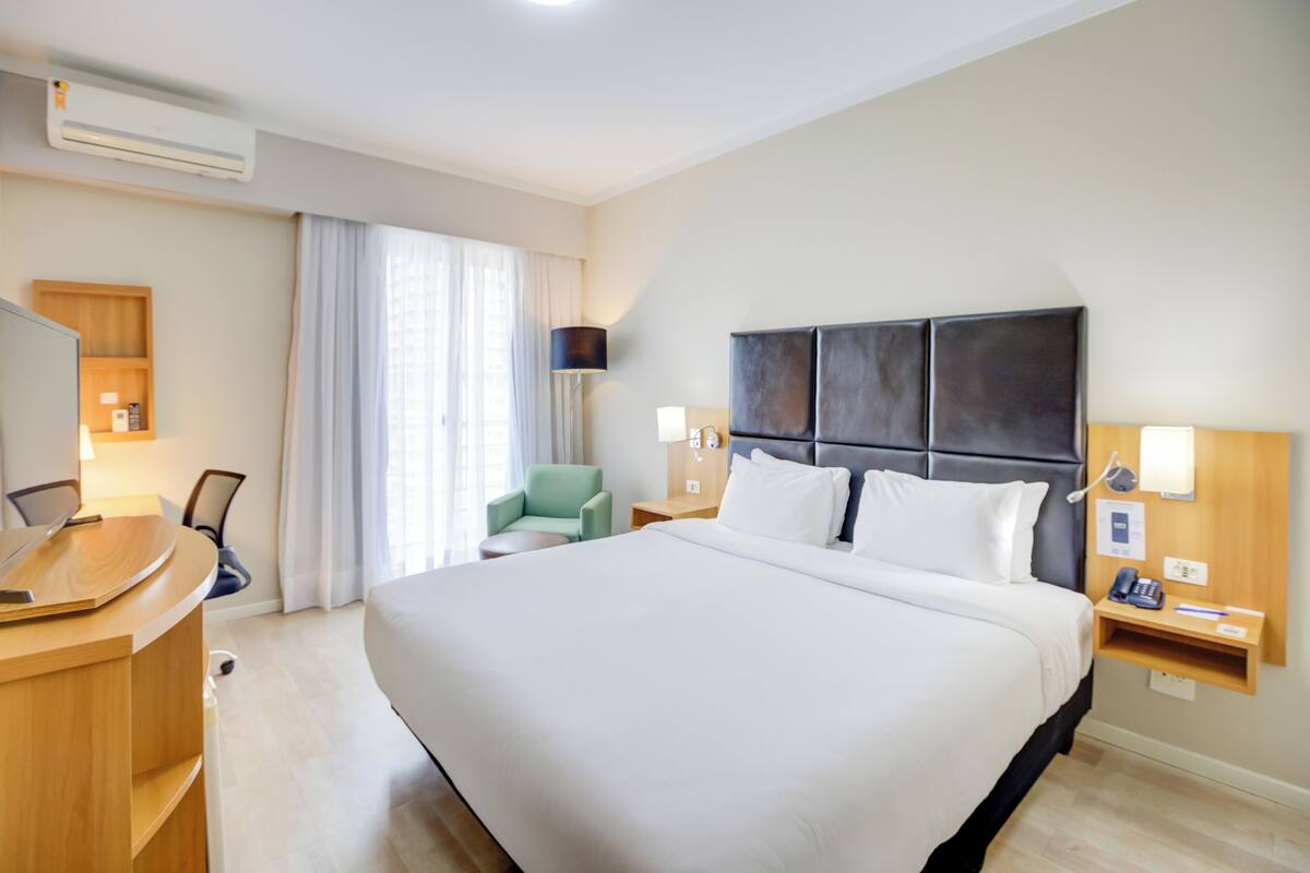 Ein Hotelzimmer mit einem großen Bett, einem Schreibtisch mit Stuhl, einem Fernseher, einer Lampe und einem Fenster mit Vorhängen.
