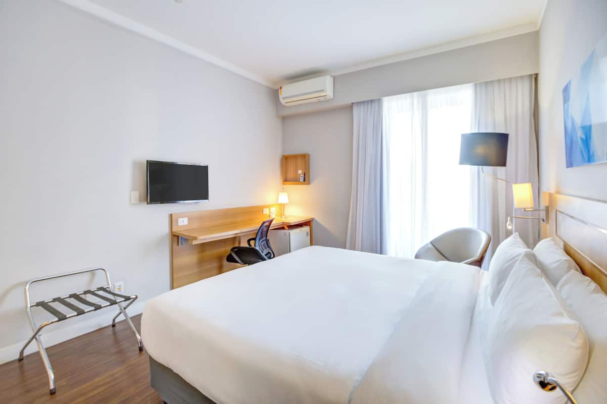 Ein Hotelzimmer mit einem Bett, einem Schreibtisch, einem Stuhl, einem Fernseher und einem Fenster mit Vorhängen.