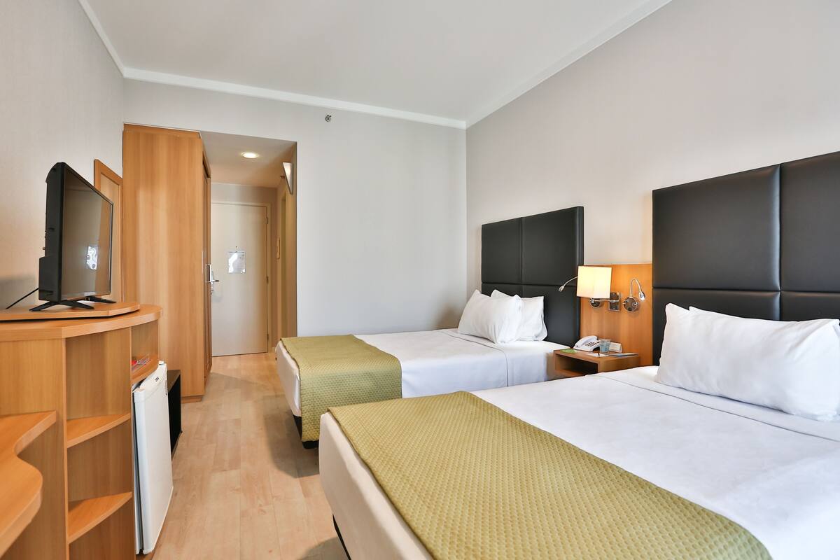 Ein Hotelzimmer mit zwei Betten, einem Schreibtisch mit Computer, einem Kühlschrank und einer Tür zu einem weiteren Raum.