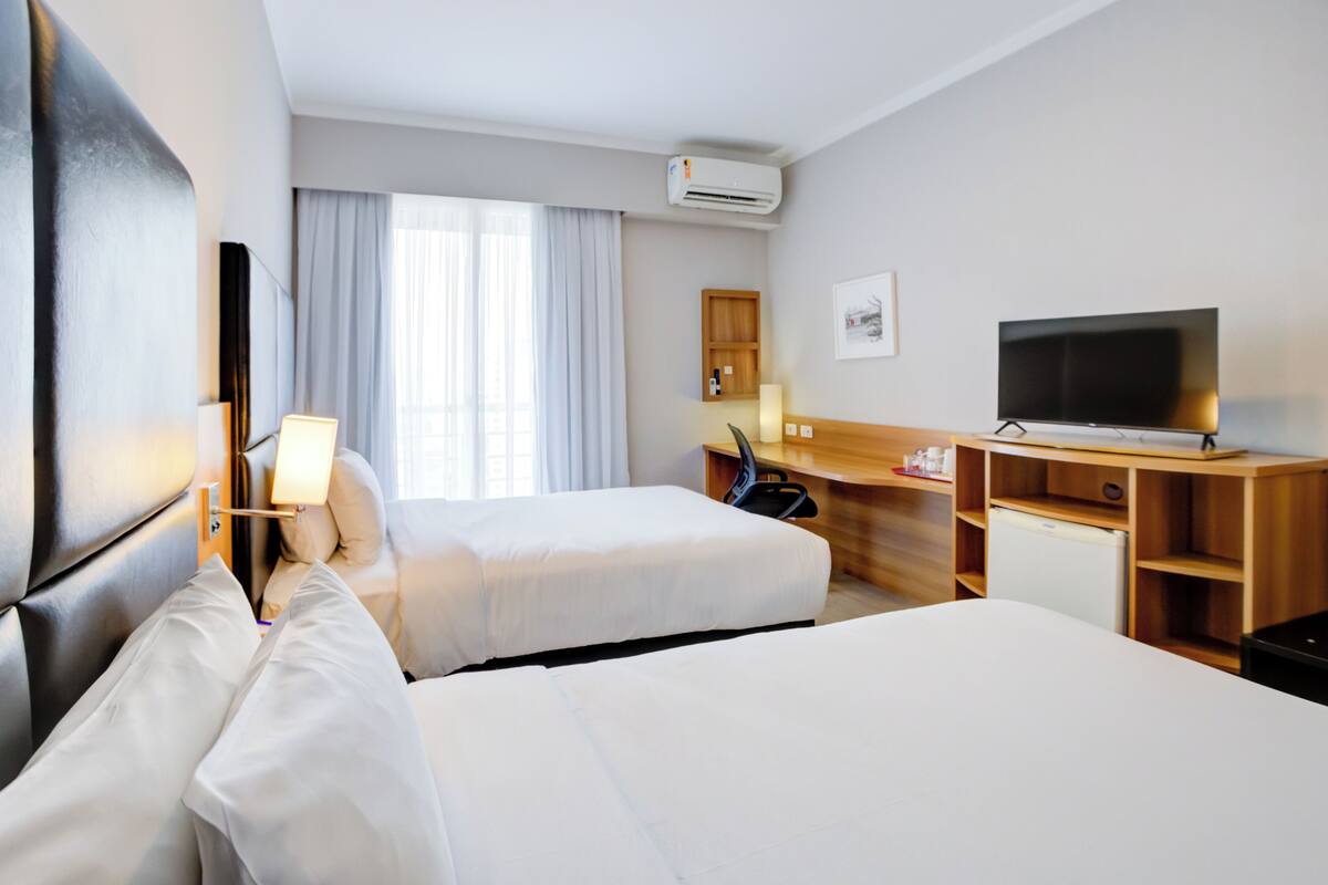 Ein Hotelzimmer mit zwei Betten, einem Schreibtisch, einem Fernseher und einer Klimaanlage.