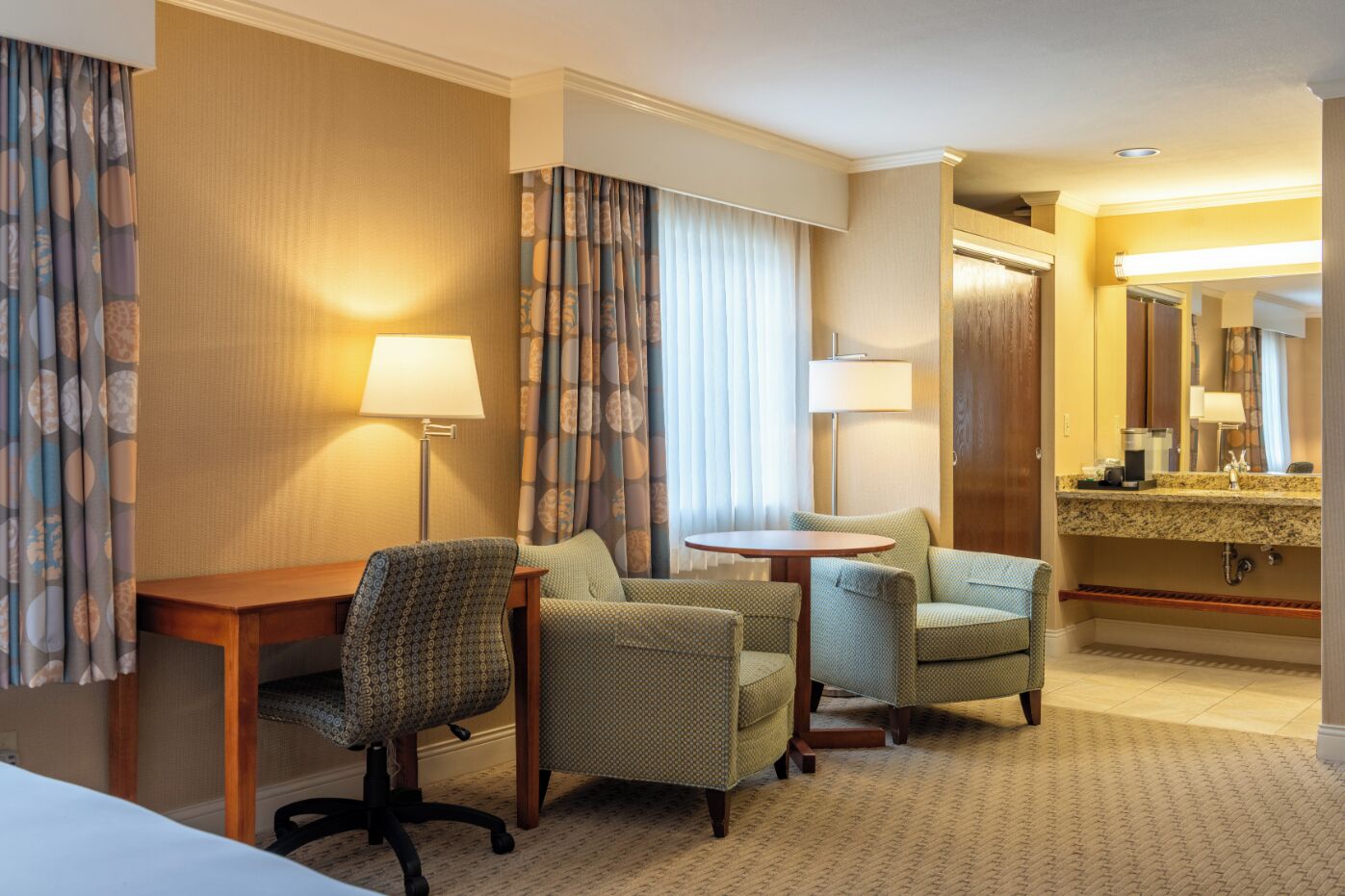 A hotel room with a desk, two armchairs, a round table, a large mirror, and a bathroom with a sink and vanity.