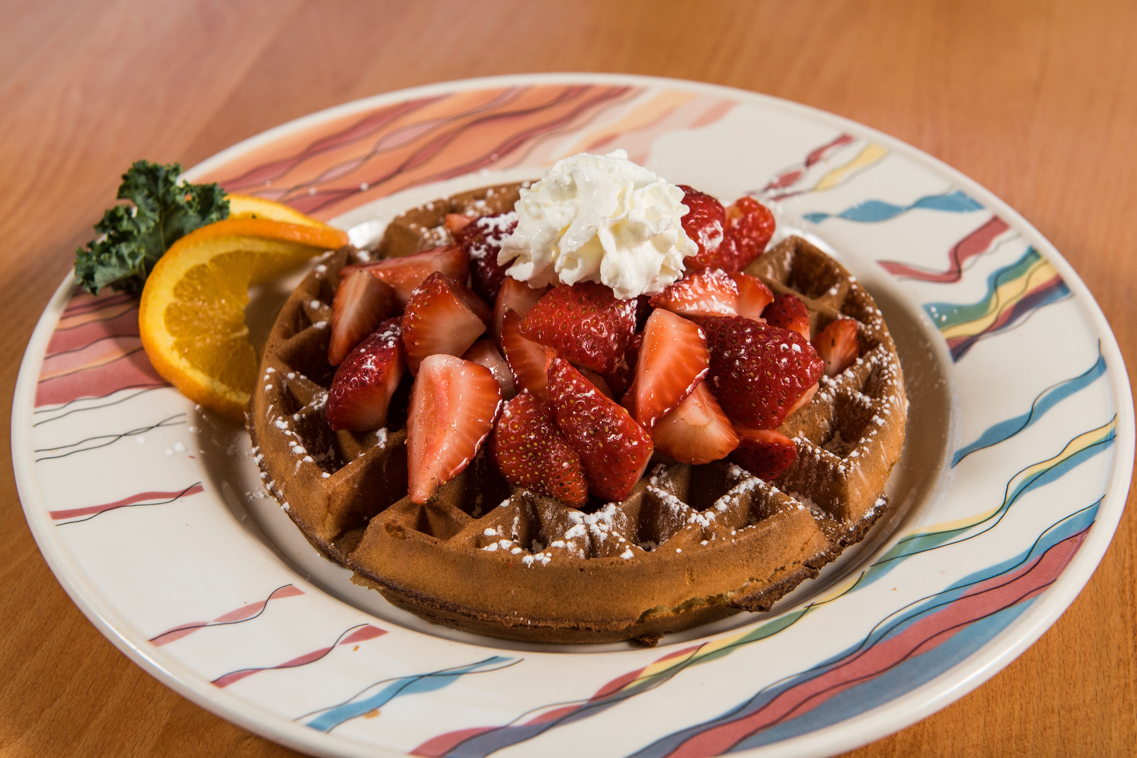Un gaufre garni de fraises, de crème fouettée et d’une tranche d’orange, le tout présenté sur une assiette colorée.
