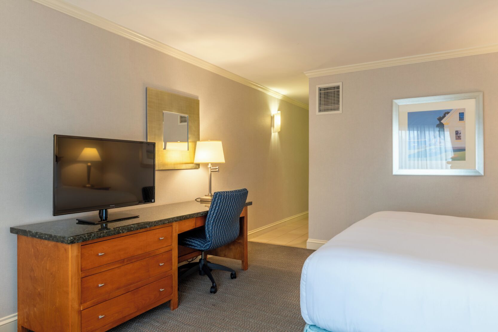 A hotel room with a bed, a desk with a television, a chair, a lamp, and a framed picture on the wall.