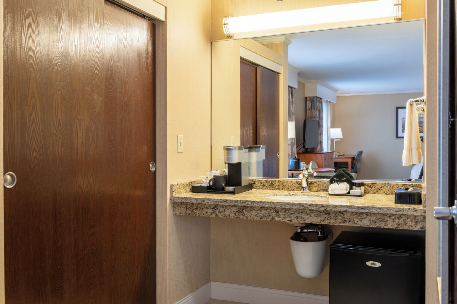 A hotel bathroom with a wooden door, a granite countertop, a sink, a mirror, a trash bin, and a black refrigerator.