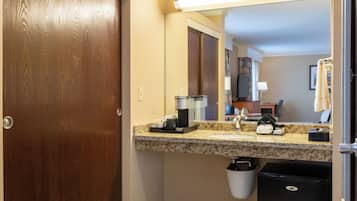 A hotel bathroom with a wooden door, a granite countertop, a sink, a mirror, a trash bin, and a black refrigerator.
