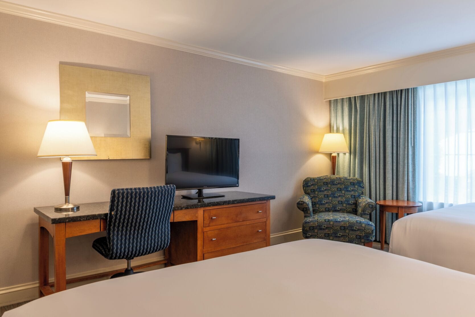 Una habitación de hotel con cama, escritorio con televisor, sillón estampado y ventana con cortinas.