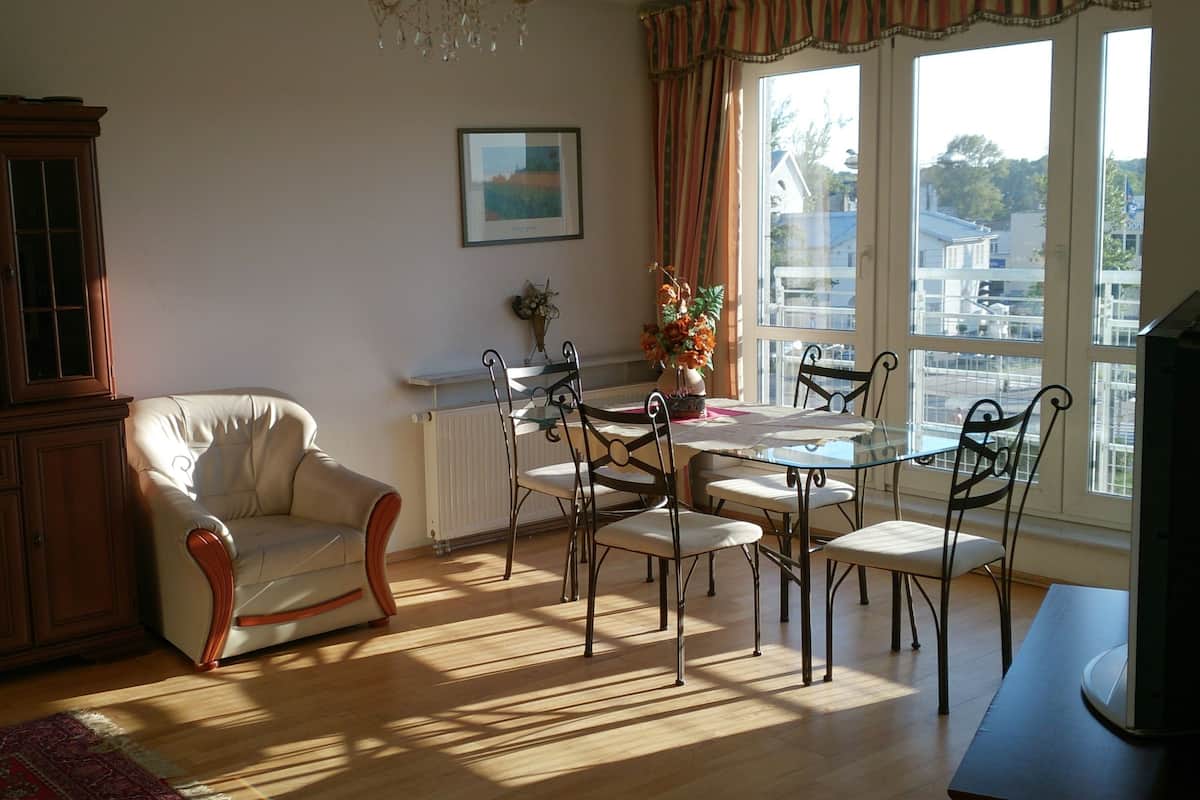 A dining area with a glass table and metal chairs, a white armchair, a wooden cabinet, and a window with curtains.