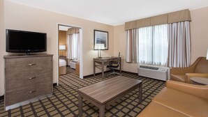 Une chambre d’hôtel équipée d’une télévision à écran plat, d’un bureau avec une chaise, d’un lit, d’un canapé et d’une grande fenêtre avec des rideaux.