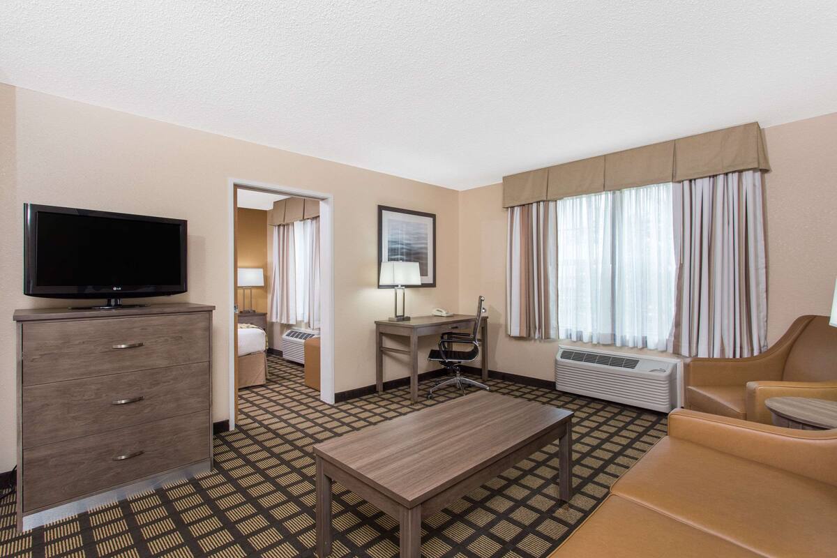 Ein Hotelzimmer mit Flachbildfernseher, Schreibtisch mit Stuhl, Bett, Sofa und einem großen Fenster mit Vorhängen.