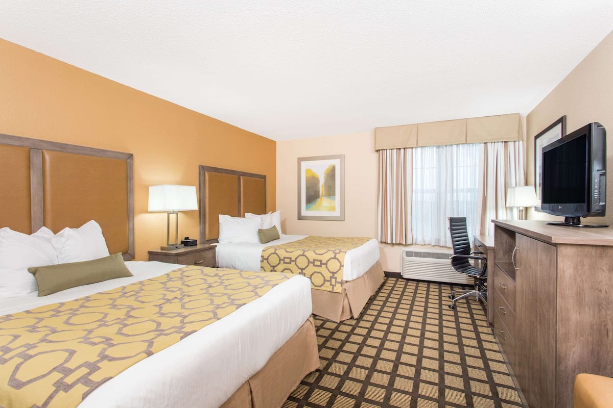 Ein Hotelzimmer mit zwei Betten, einem Schreibtisch mit Computer, einem Fernseher und einem Fenster mit Vorhängen.