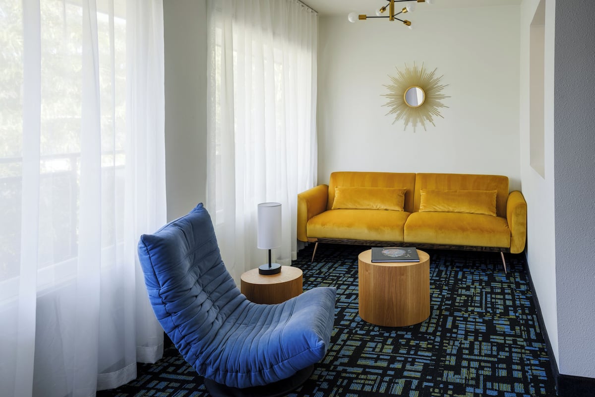 A modern living room with a blue chair, a yellow sofa, and a sunburst mirror on the wall.