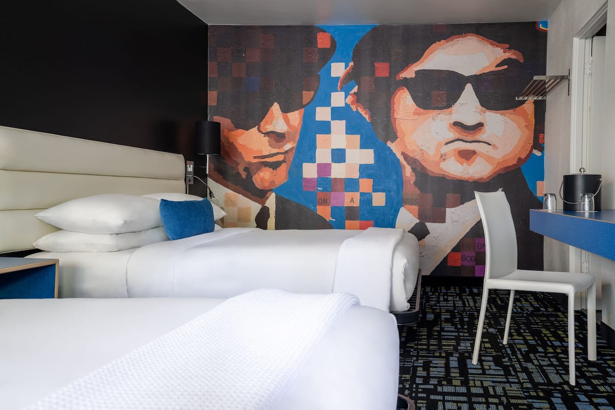 A hotel room with a bed, a desk, a chair, and a large pixelated mural on the wall.