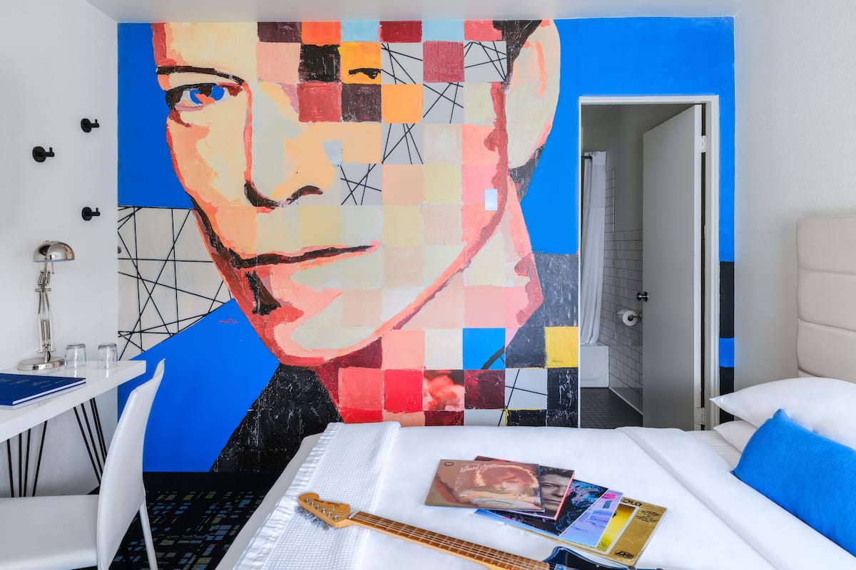 A hotel room with a large abstract painting on the wall, a bed with a blue guitar and music albums, and a white desk with a lamp.