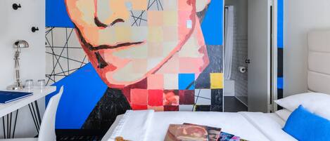 A hotel room with a large abstract painting on the wall, a bed with a blue guitar and music albums, and a white desk with a lamp.