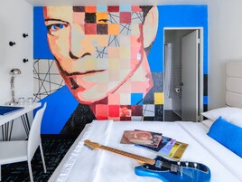 Una habitación de hotel con un cuadro abstracto grande en la pared, una cama con una guitarra azul y discos de música, y un escritorio blanco con una lámpara.