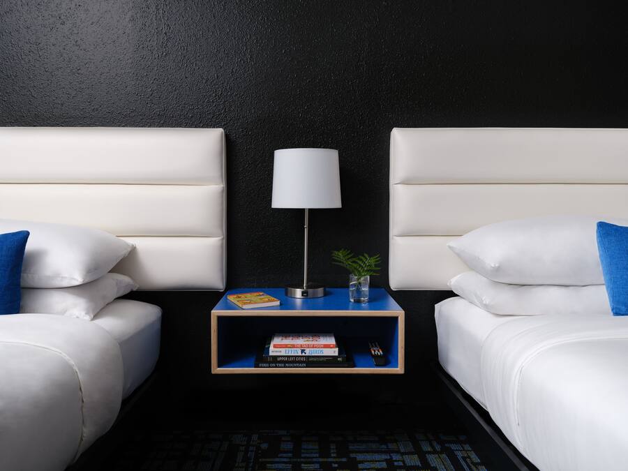 Una habitación de hotel con dos camas, una mesita de noche con una lámpara, una planta pequeña y una pila de libros.