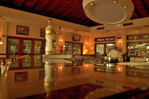 Um saguão de hotel com uma receção em mármore polido, uma grande fonte decorativa e uma televisão na parede.