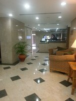 Um hall de entrada com balcão de check-in, mesa de recepção, uma planta grande em vaso, sofá de vime e piso revestido.