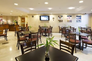 Uma sala de jantar de hotel com várias mesas e cadeiras, uma mesa de recepção e uma televisão na parede.